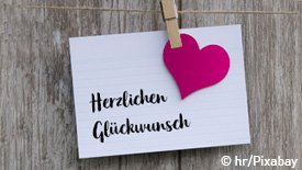 Ein Zettel ist mit einer Klammer an einer Leine vor einer Holzwand befestigt. Auf dem Zettel steht "Herzlichen Glückwunsch" und es ist ein Herz im Farbton Fuchsia in der rechten Hälfte des Zettels.