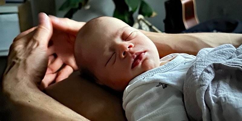 EIn Baby schläft auf dem Schoß seines Vaters, dessen Hände um den Kopf des Kindes gelegt sind