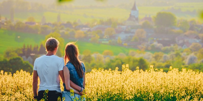 Rückenansicht zweier Menschen, die Arm in Arm vor einem Rapsfeld stehen und in die Weite schauen