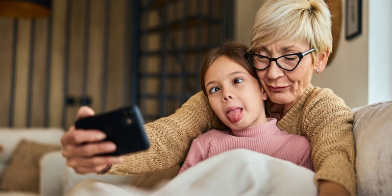 Oma und Enkelin auf einer Couch machen ein Selfie, das Mädchen schielt und streckt seine Zunge raus