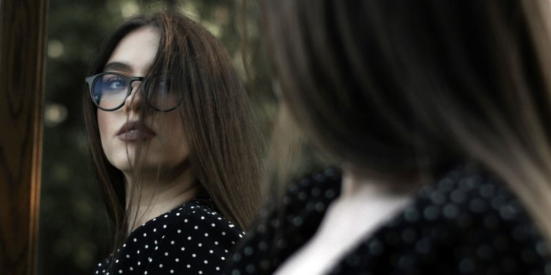 junge Frau mit Brille und langen Haaren schaut ernst in einen Spiegel