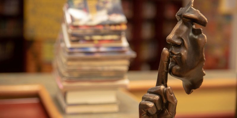 Seitenansicht einer Bronze-Maske, die den Zeigefinger vor den Mund legt, dahinter ein Stapel Bücher