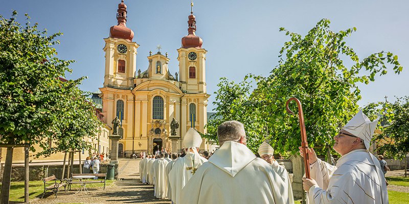 Die gelbe Wallfahrtskirche "Maria Heimsuchung" im Sonnenlicht, im Vordergrund eine Prozession von Geistlichen  