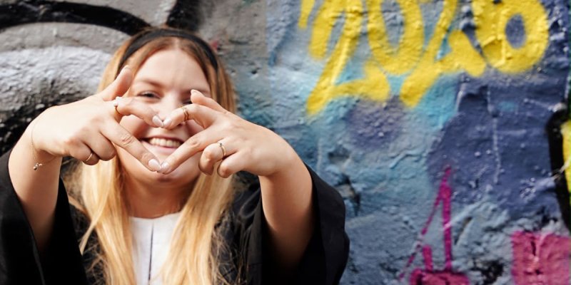 eine junge Pfarrerin vor einer Wand mit Graffiti formt mit ihren Händen ein Herz