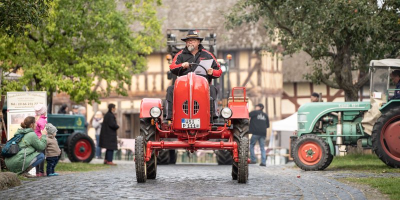 Mann mit Hut auf einem Oldtimer-Traktor im Freilichtmuseum Hessenpark mit alten Fachwerkhäusern, am Rande Zuschauer