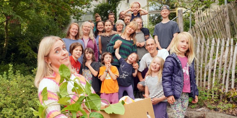 Reporterin Nina Fehr mit Umzugskiste vor einer Gruppe von 20 lachenden Personen unterschiedlichen Alters, die in einem Garten stehen